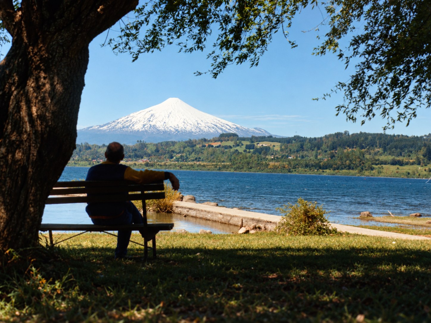 Hombre sentado en un banco frente a un lago, con un volcán nevado al fondo